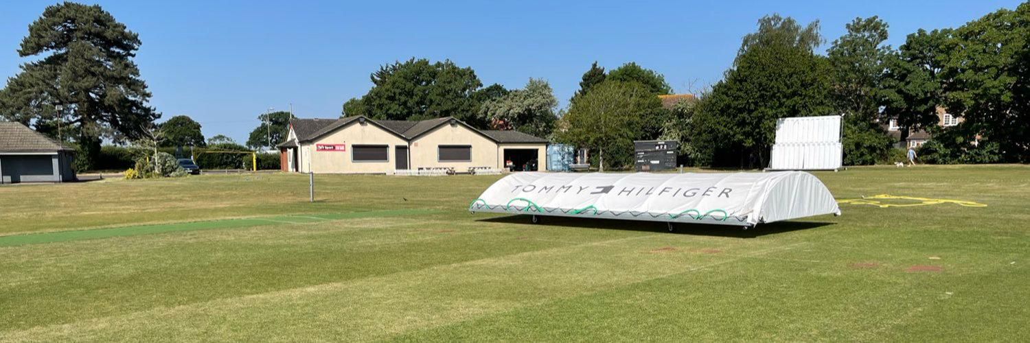 Image of a cricket pitch on a lovely sunny day with the covers on the strip and the sports and social club house in the background
