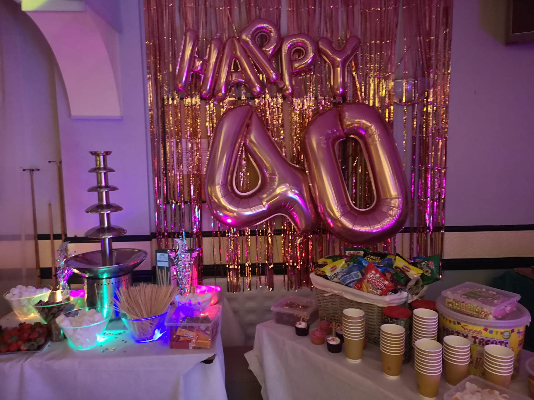 Picture shows two buffet tables all laid ready for a party. The contain a selection of party foods, cups, plates and a chocolate fountain. In the background you can see a gold, tinsel backdrop with Happy 40 balloons.