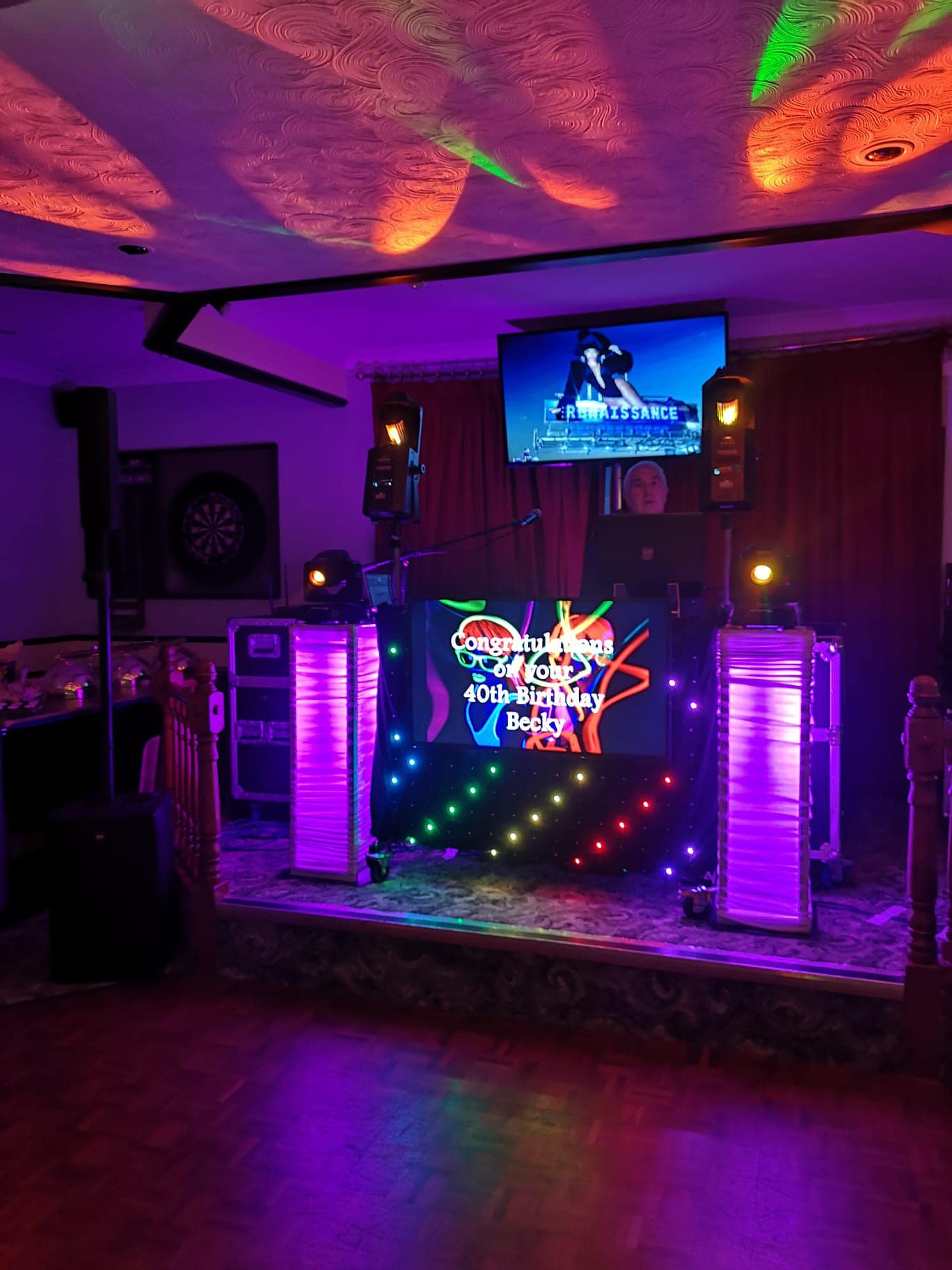 Picture shows a DJ desk with speakers and lights on a raised platform. The front of the DJ stand displays a Happy Birthday message. You can see the disco lights reflecting on the ceiling and on the dancefloor.