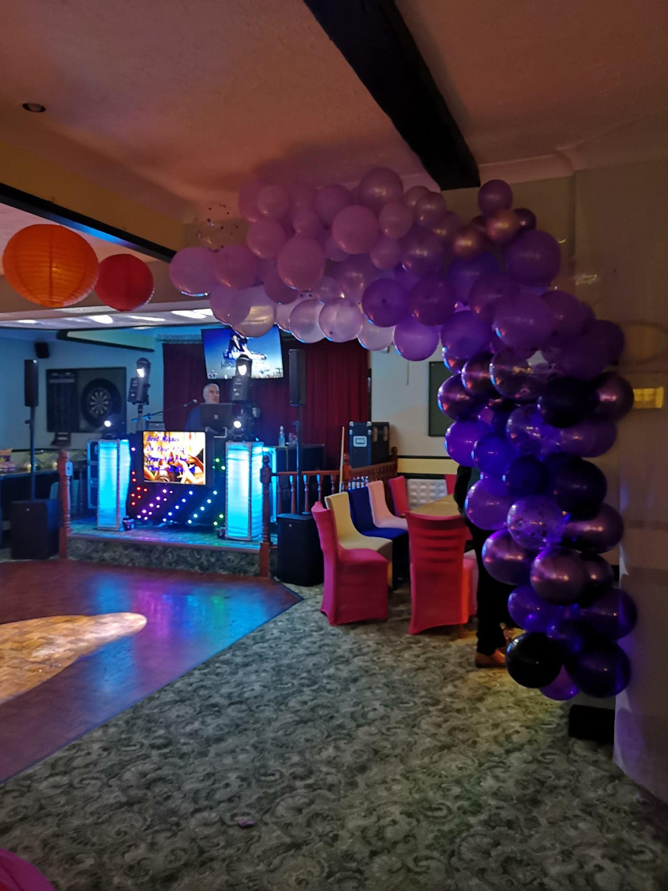 Picture shows a function room with lovely lilac and purple balloon arch ready for a party. In the background you can see the DJ stand all lit-up with disco lights, the dancefloor and a table and chairs.