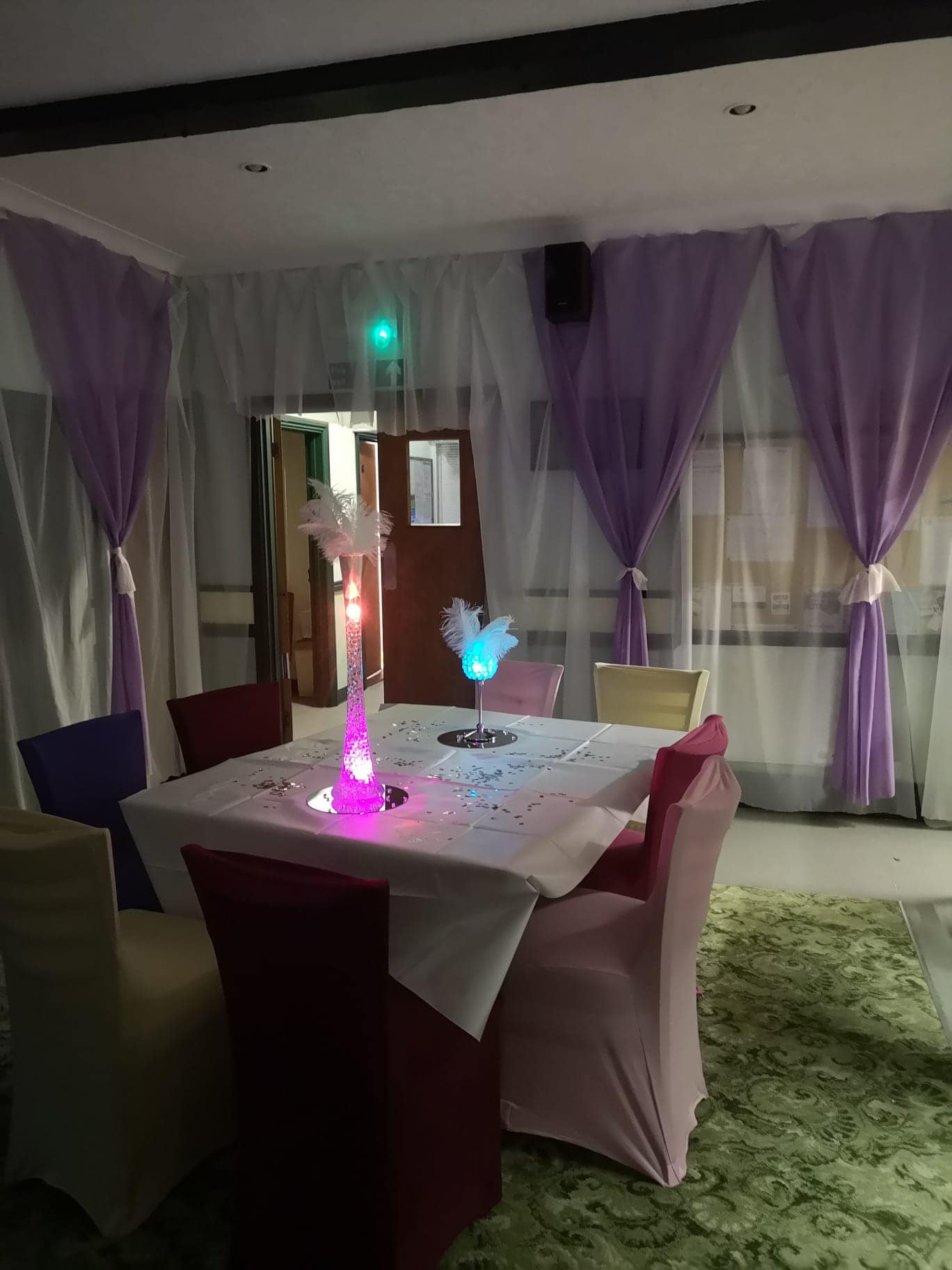 Picture shows a nicely laid table with a tall, thin, lit-up table decoration ready for a party. In the background there are white and lilac chiffon curtains which match the party decorations.